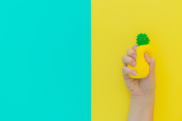 Flat lay antistress toy squish yellow pineapple with green leaves in hand.Bright blue background.Compressing, soft, squeezable items to relieve stress, problems, anxieties, worries. Summer concept