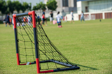 Obraz premium A small size of black and red plastic goal with black net for junior football teams placed on a school tournament pitch. Seeing groups of players and parents walking around in a background.
