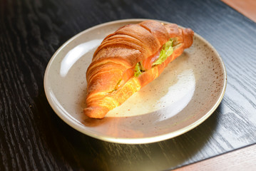Fresh baked croissant filled with ham and lettuce served on white plate in restaurant or cafe.