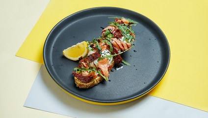 Close up view on Appetizing toast on rye bread with arugula, poached egg, salmon and sun-dried tomatoes on a black plate on colored background. Breakfast food