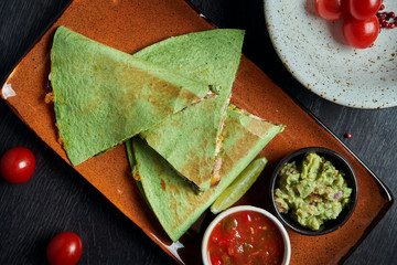 Appetizing baked quesadilla with minced chicken of cheese in green tortilla on a ceramic plate on a black background. Modern mexican cuisine.
