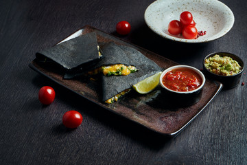 Appetizing baked quesadilla with 4 types of cheese in black tortilla on a ceramic plate on a black background. Modern mexican cuisine.