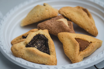 Hamantaschen. (ozen haman). Jewish food for Purim. Triangular crispy dough, stuffed with dates, halva and chocolate, on a white plate.