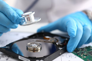 A male repairman wearing blue gloves holding stethoscope on hard drive from computer laptop in...