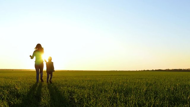 A mother and child hold hands as they run through the green grass at sunset. The concept of a happy family, travel and health.