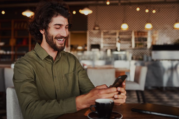 Cheerful handsome young man relaxing in modern cafe using cellphone while drinking coffee