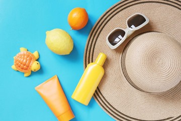 Flat lay summer photo. Beach essentials on a blue background. Yellow sunscreen tube, body cream, sun hat, sunglasses, fruits and toy turtle