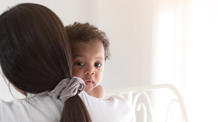Asian mom holding adorable mixed race African baby boy on the sholder with white curtain as copy...