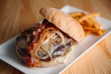 meat burger with bacon, onion rings and french fries in a white plate on a wooden table