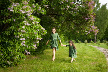 Fototapeta premium Two sisters in green linen dress have fun in the park with blooming lilacs, enjoy spring and warmth
