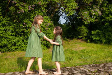 Naklejka na ściany i meble Two sisters in green linen dress have fun in the park with blooming lilacs, enjoy spring and warmth