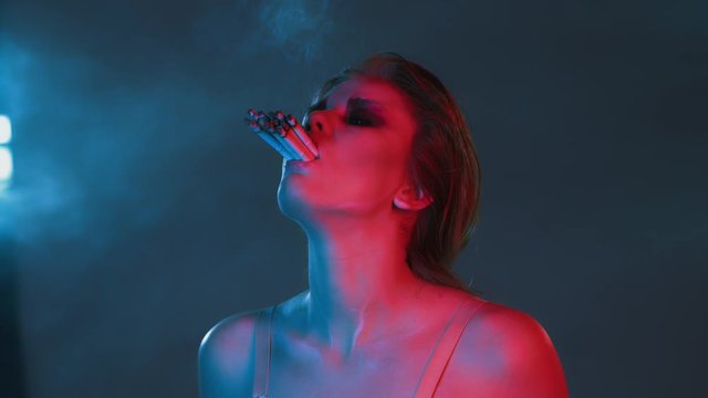 young beautiful girl put a lot of cigarettes in her mouth, smoke everywhere, personification of excessive use and addiction on nicotine and cigarette, neon blue and red light