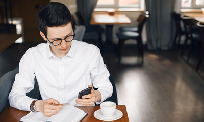Handsome caucasian man wearing glasses is having a break with a hot coffee in a restaurant while texting and holding a bank card