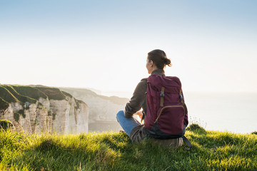 Young woman traveler with backpack sitting and looking at sea in Normandy, France over beautiful cliffs background, Travel, active lifestyle and summer holiday concept - obrazy, fototapety, plakaty