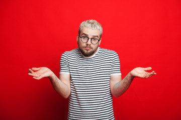 Handsome caucasian boy with beard and blonde hair is looking through eyeglasses and showing his indecisiveness on a red background - obrazy, fototapety, plakaty