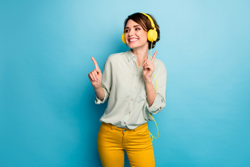 Photo of funny cheerful lady listening radio cool stylish earphones raise fingers discotheque dj dancing wear casual green shirt yellow trousers isolated blue color background