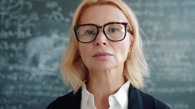 Portrait of serious adult woman scientist in glasses looking at camera in university class standing against blackboard with formulas. People and science concept.