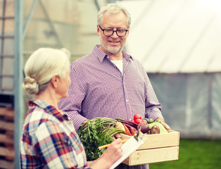 farming, gardening, agriculture, harvesting and people concept - senior couple with box of vegetables and clipboard at farm greenhouse