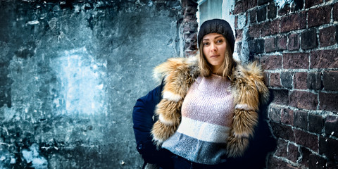 Image of a young beautiful girl dressed in a winter jacket with a fur collar on a brick wall background of an old building. The photo contains artistic noise, selective focus.