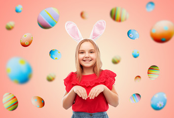 easter, holidays and childhood concept - happy girl wearing bunny ears headband over colored eggs on living coral background