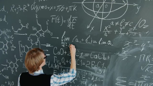 High angle view of mathematics teacher handsome young man in glasses writing formulas and equations on board with chalk. People and education concept.