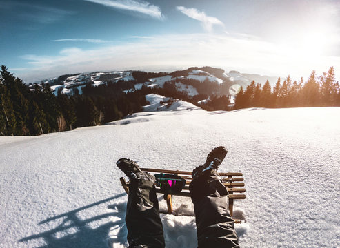 Pov View Of Young Man Looking The Sunset On Snow High Mountains With Vintage Sledding - Legs View Of Travel Influencer Enjoying Winter Landscape - Vacation Concept - Focus On Wood Luge