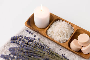 Obraz na płótnie Canvas beauty and wellness concept - sea salt, soap and candle on wooden tray and bunch of lavender on bath towel