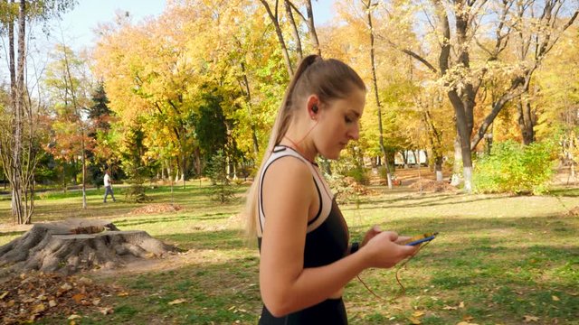 4k video of beautiful young woman with ponytail setting music plaer on her smartphone and starting to run during fitness exercise in autumn park