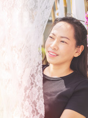 Happy Thai asian woman wear black t-shirt is smiling, peeking back the white soft lace fabric curtain on sunshine summer day. Asia Lifestyle outdoor.