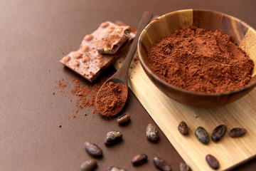 sweets, confectionery and culinary concept - chocolate with hazelnuts, cocoa beans and powder in wooden bowl with spoon on brown background