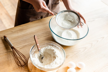 cropped view of young woman sieving flour in glass bowl near whisk, spoon and eggs