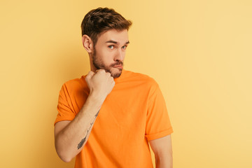 angry young man threatening while holding fist near jaw on yellow background