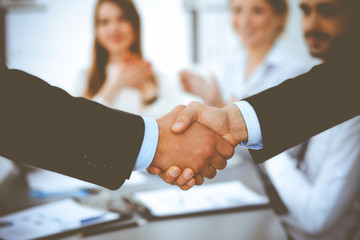 Business people shaking hands at meeting while theirs colleagues clapping and applauding. Group of unknown businessmen and women in modern white office. Success teamwork, partnership and handshake