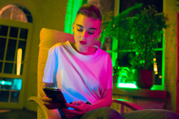 Beautiful. Cinematic portrait of stylish woman in neon lighted interior. Toned like cinema effects, bright neoned colors. Caucasian model using smartphone in colorful lights indoors. Youth culture.