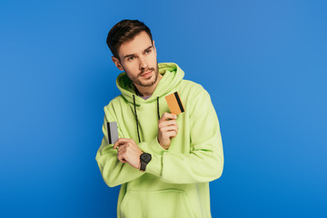 pensive young man looking away while holding credit cards on blue background