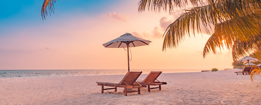 Best beach sunset panorama. Tropical landscape scene. Amazing sunset sea view with palm leaves and two lounge chairs with umbrella. Luxury lifestyle, travel and summer vacation background