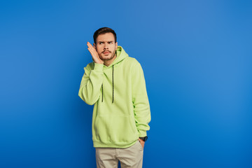 curious young man holding hand near ear while looking away isolated on blue