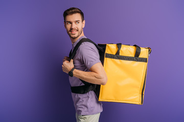 cheerful delivery man looking away while carrying thermo backpack on purple background