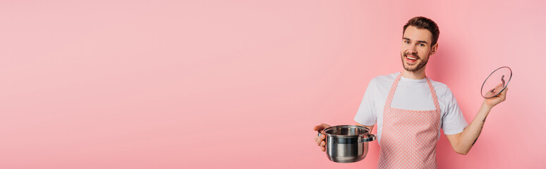 panoramic shot of happy young man in apron opening saucepan on pink background