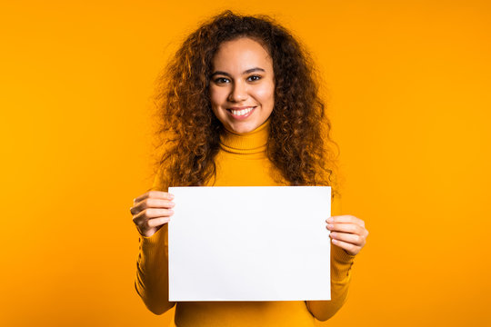 Pretty curly woman holding horizontal white a4 paper poster. Copy space. Smiling trendy girl on yellow background.