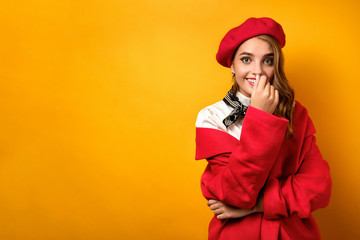A beautiful girl in a red coat and beret, with a neckerchief stands on a yellow background and laughs hiding behind her hand