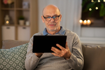 technology, old age and people concept - happy bald senior man with tablet pc computer sitting on sofa at home in evening