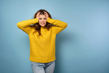 A brunette in a yellow sweater stands on a blue background, frowning and ruffling her hair, looking into the frame.