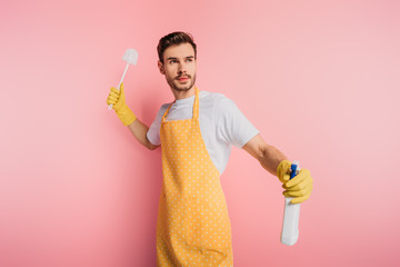 angry young man in apron and rubber gloves looking away while holding spray bottle and plunger on pink background