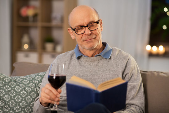 old age, leisure and people concept - happy senior man with glass of red wine reading book at home in evening