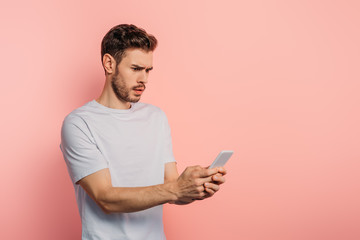 shocked young man chatting on smartphone on pink background