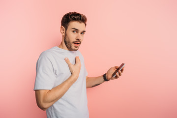 shocked young man touching chest while holding smartphone on pink background