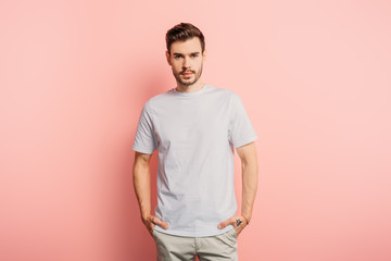 handsome, confident young man with hands in pockets looking at camera on pink background