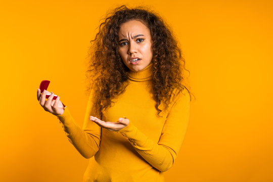 Disappointed Upset Girl, She Doesn't Like Gift. Pretty Mixed Race Young Woman In Yellow Holding Small Jewelry Box With Proposal Diamond Ring On Studio Background.