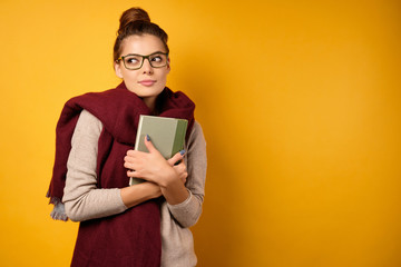 A beautiful brunette with a high bun and glasses stands in a sweater and scarf, presses a book to her chest, looks to the side